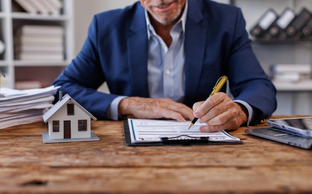 Signature d'un contrat immobilier par un professionnel Agent immobilier en costume signant un contrat de vente à côté d'une maquette de maison sur un bureau en bois.