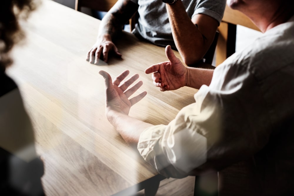 Communication et réunion d'équipe autour d'une table Discussion professionnelle animée avec gestuelle des mains autour d'une table en bois ensoleillée.