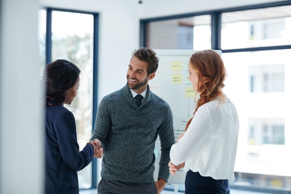 Collaboration et poignée de main entre collègues au bureau Trois professionnels souriants se serrent la main devant un tableau blanc dans un bureau moderne et lumineux.